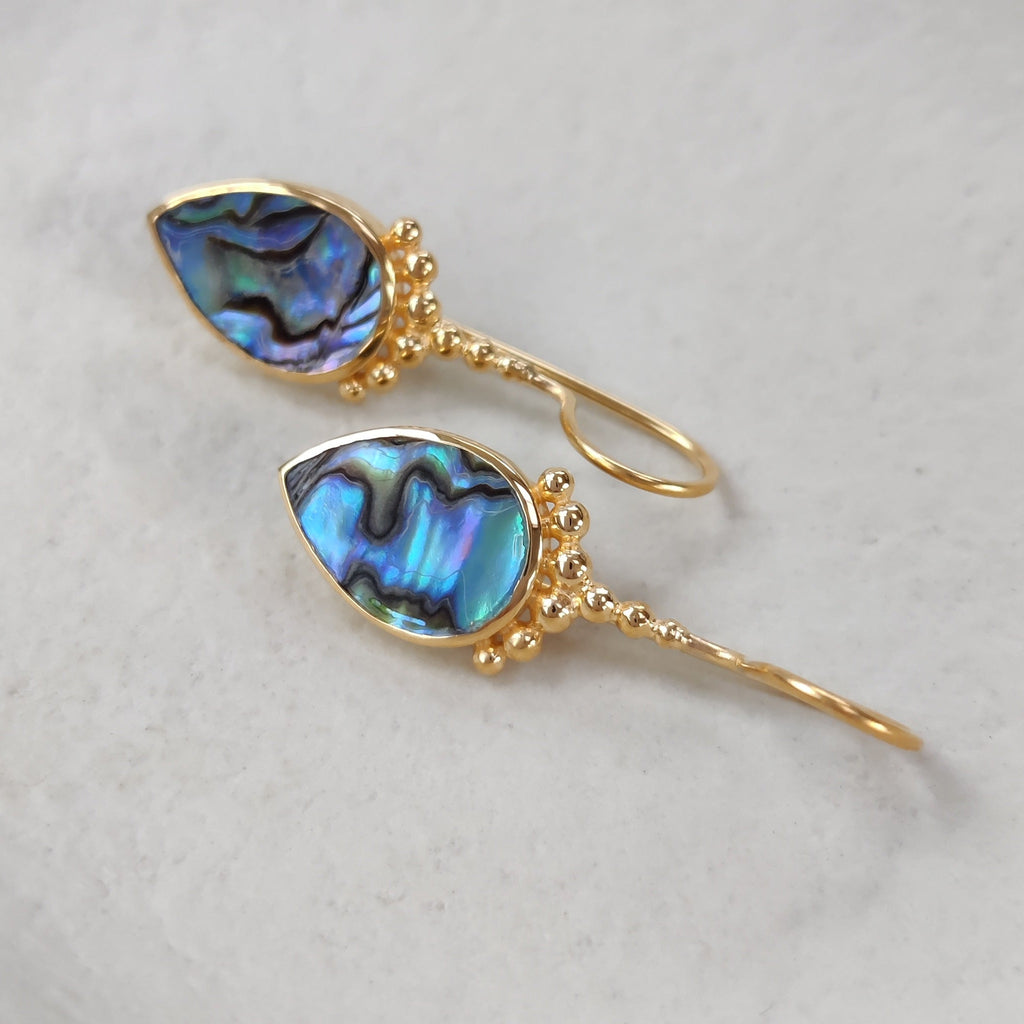 Drop-shaped gold earrings with shimmering blue abalone shells on a white dish