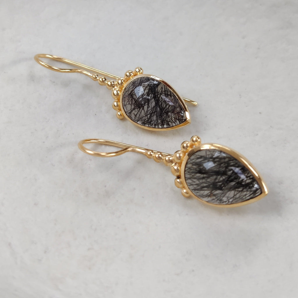 Drop-shaped gold earrings with black rutile stones on a white dish