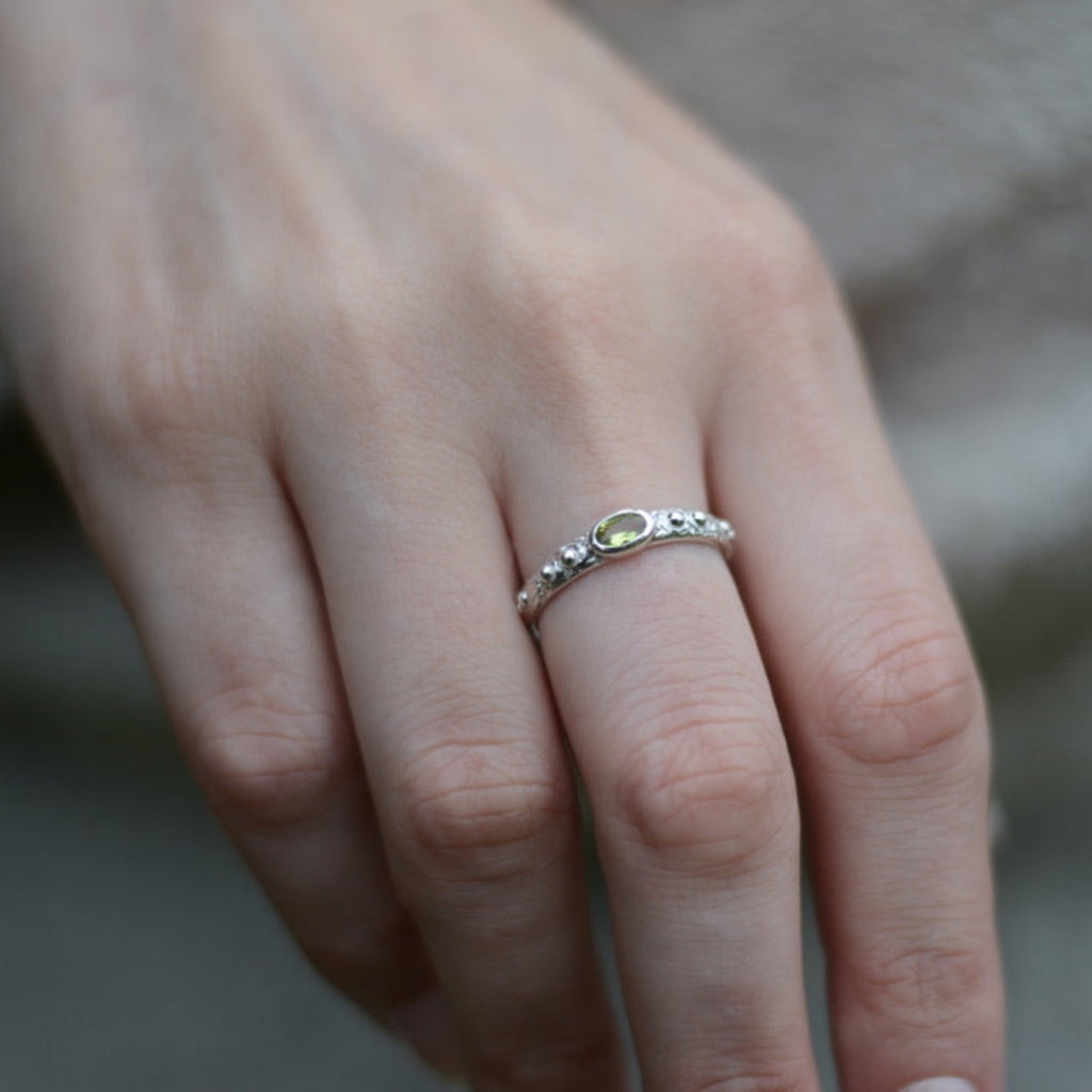 Close-up of a hand wearing a silver ring with a faceted green gemstone on a neutral background.