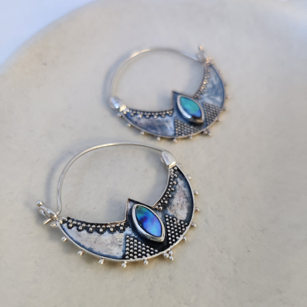 Close-up of silver crescent hoop earrings featuring shimmering abalone shell, highlighting their intricate design and iridescent colors.