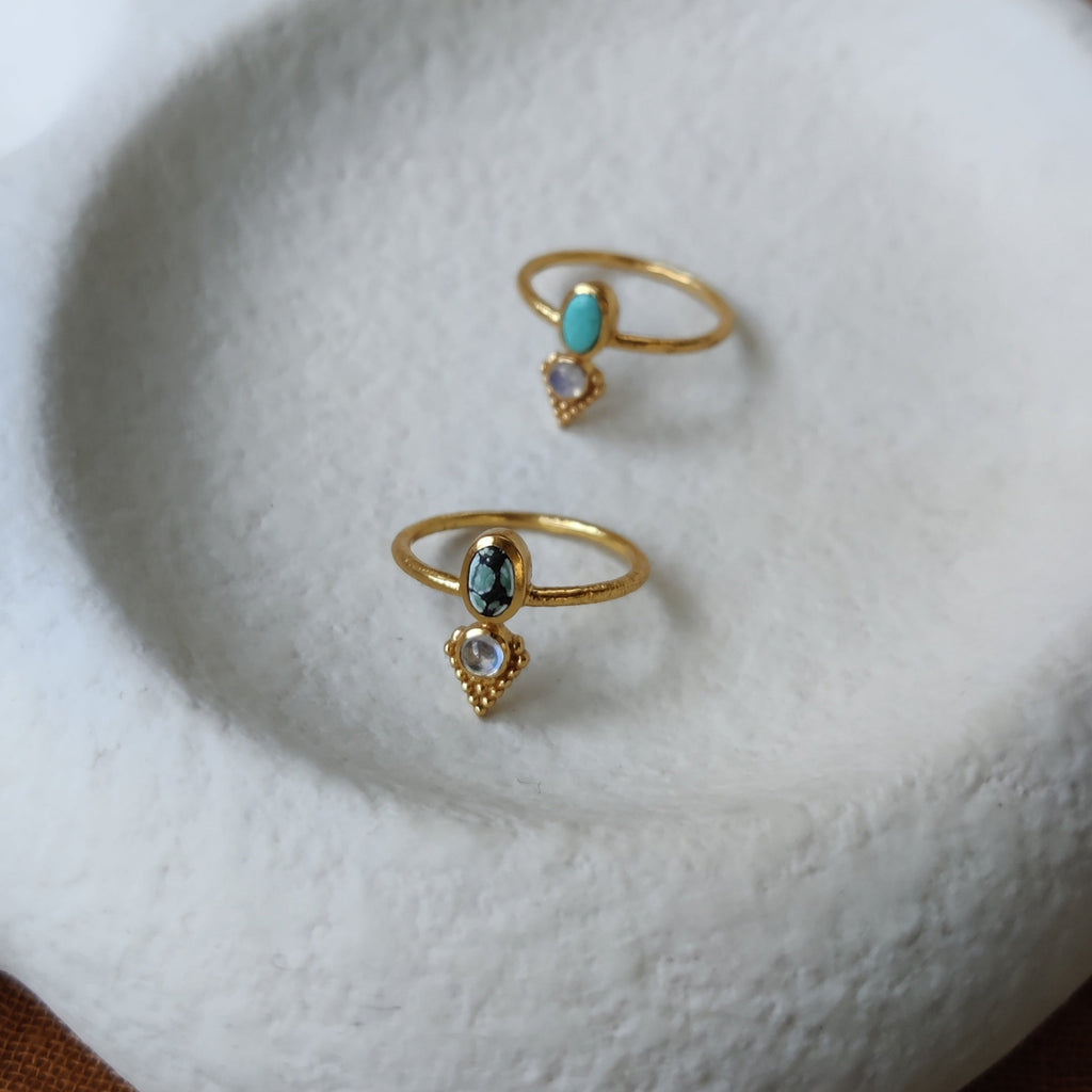 Dainty brass rings with turquoise and moonstone, displayed on a jewelry plate.