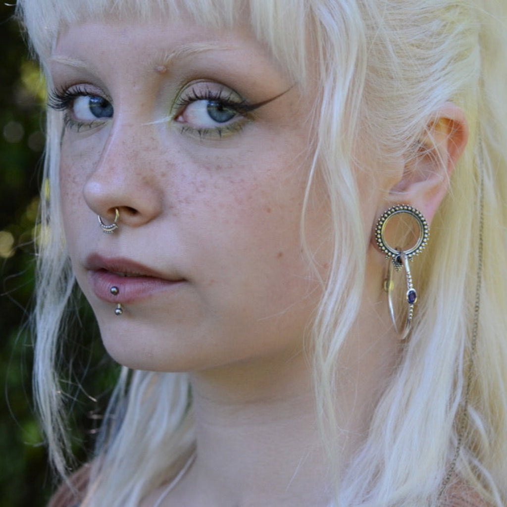 Person with blonde hair and piercings, wearing large hoop earrings against a blurred natural background