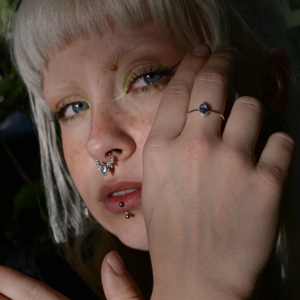 Person with multiple piercings holding her hand  with a tanzanite silver ring near the face against a dark background