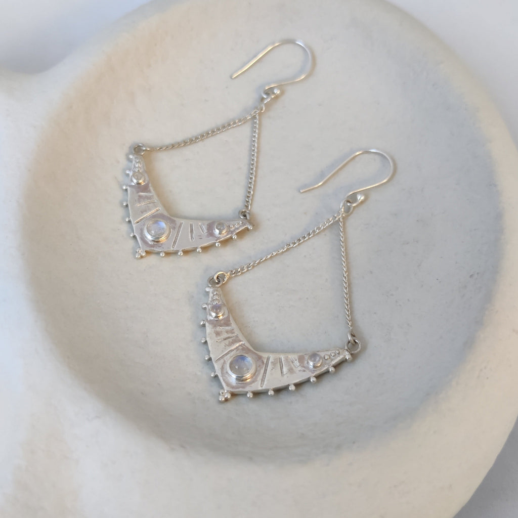 Dangling silver earrings with three moonstones, displayed on a jewelry plate.