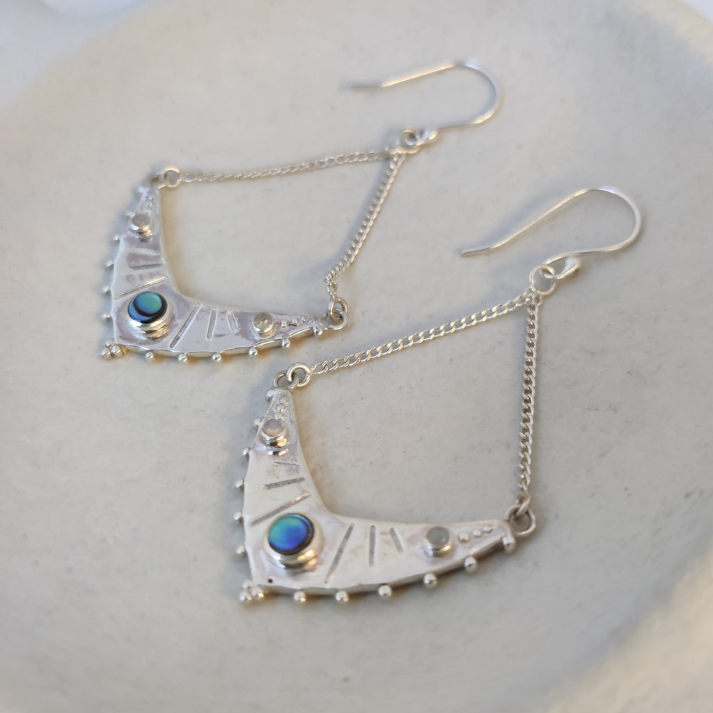 Dangling silver earrings with abalone shell and moonstone, displayed on a jewelry plate.