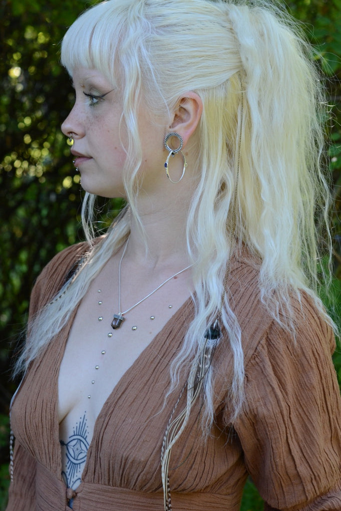 Woman with long blonde hair styled in a high ponytail, wearing silver jewelry and a brown dress outdoors.