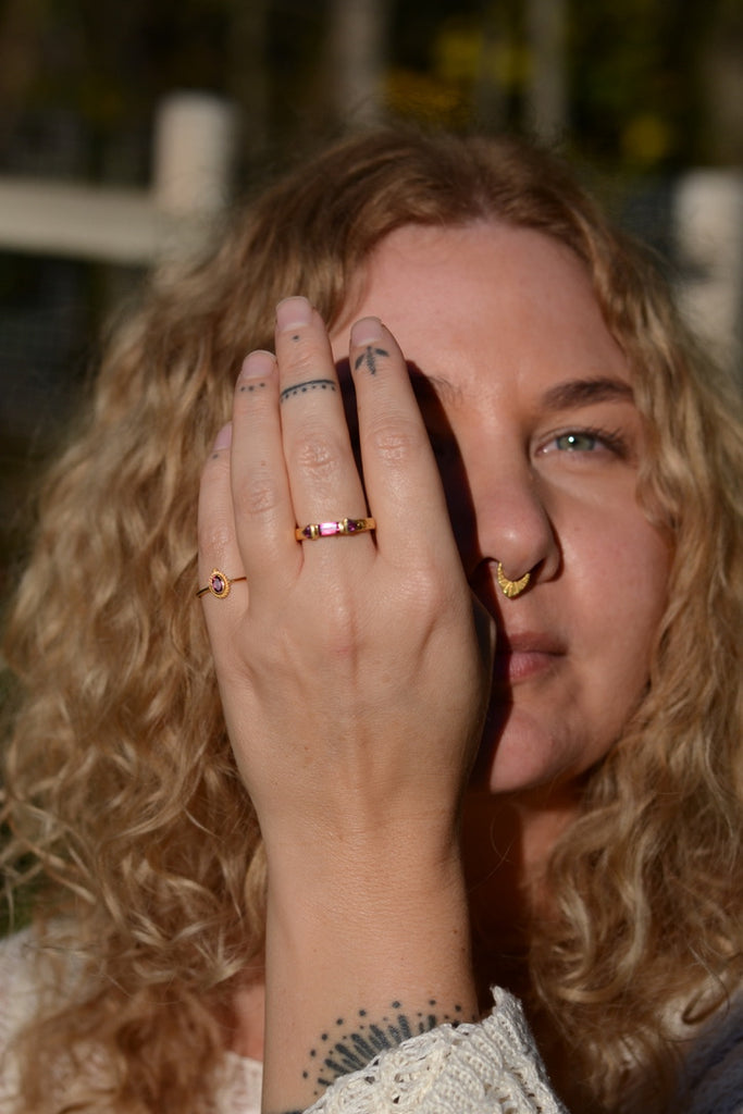 Woman with curly hair holding hand near face, wearing multiple gold rings with rhodolite garnet and a nose ring.