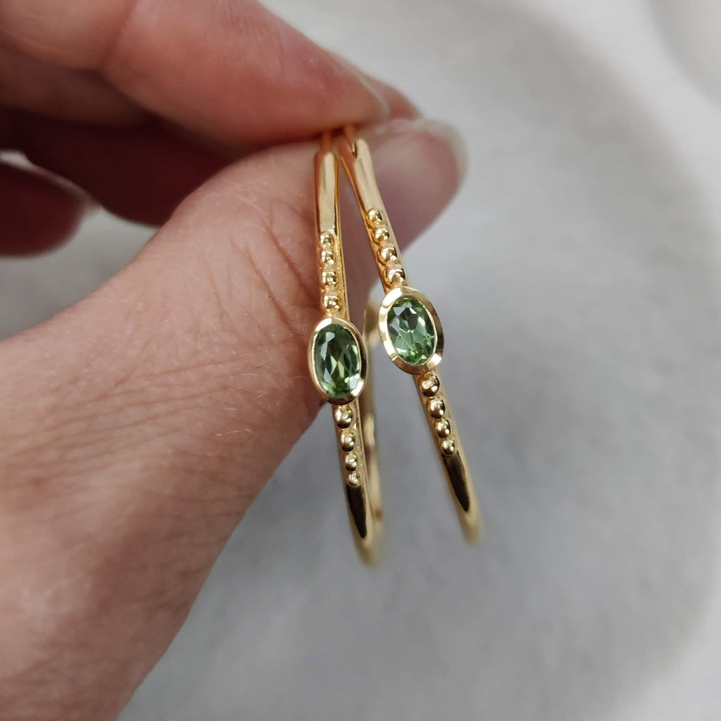 Gold hoop earrings with green gemstones held by a hand against a neutral background
