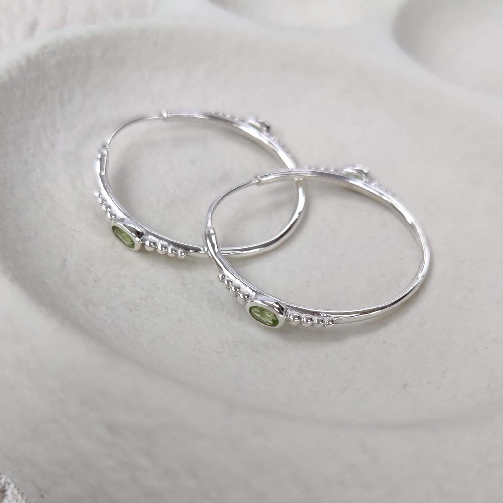 Bohemian silver hoop earrings with faceted green gemstones and dotted details on a white surface.