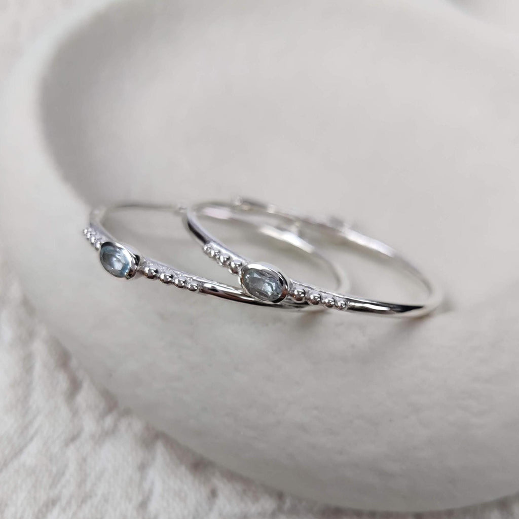 A pair of bohemian silver hoop earrings with faceted blue topaz gemstones on a white surface.