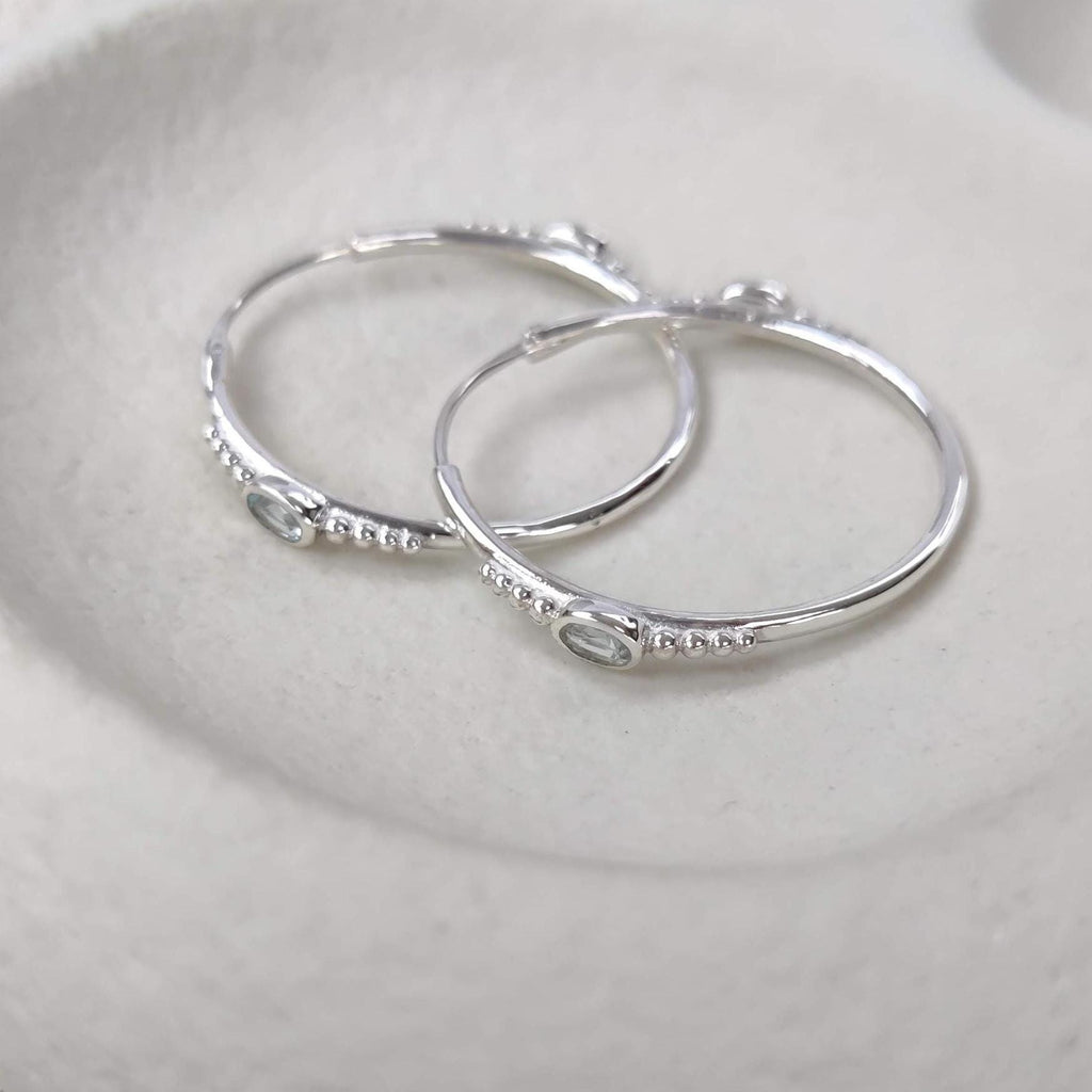 Bohemian silver hoop earrings with faceted blue topaz gemstones on a white surface.