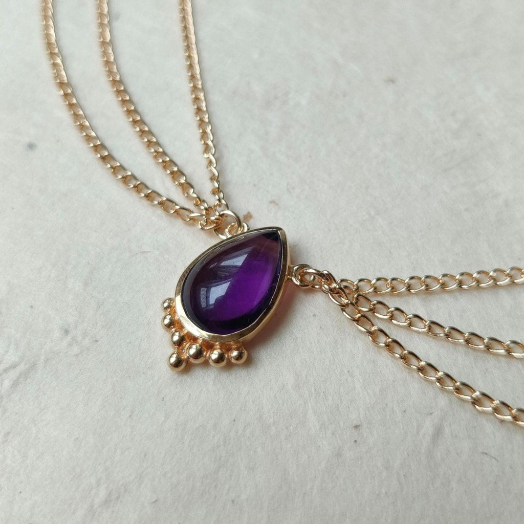 Gold head chain with drop-shaped purple amethyst gemstone centerpiece, and three chains draping on each side.