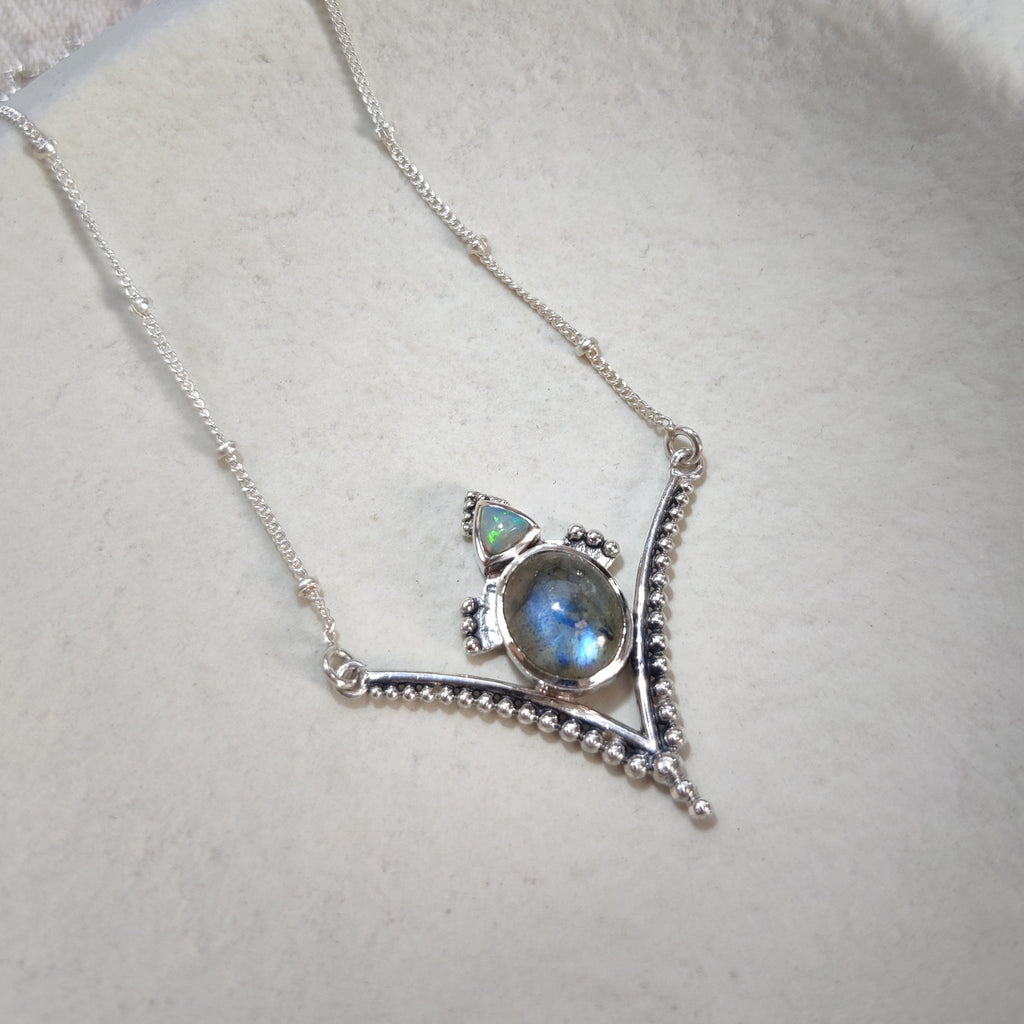 A V-shaped silver pendant with a blue labradorite gemstone and a sparkling opal, displayed on a white jewelry plate.