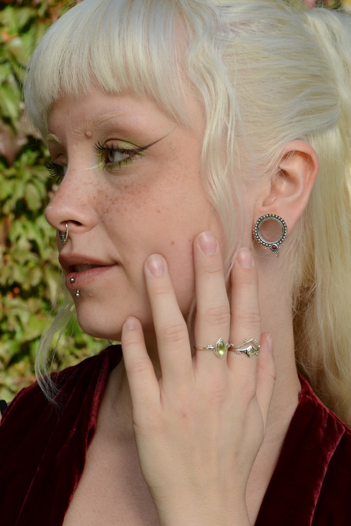 Person with blonde hair and piercings, wearing silver rings with peridot, against a blurred green background