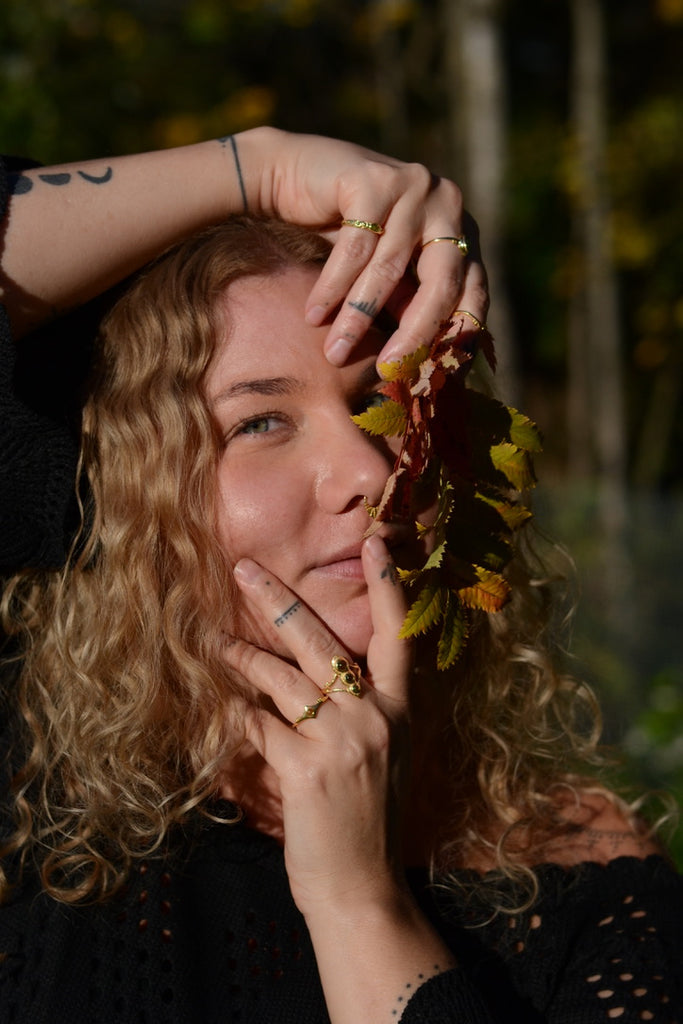 Woman with curly hair holding autumn leaves near her face outdoors, wearing gold rings with green gemstones