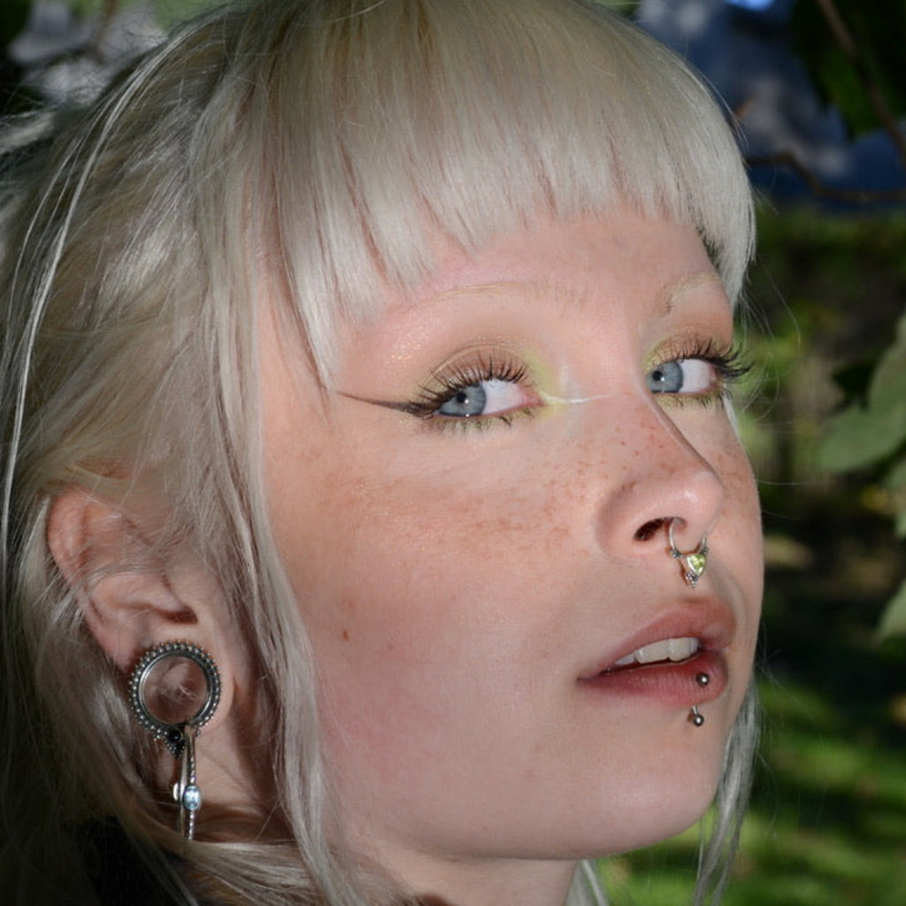 Close-up of a person with platinum blonde hair and piercings, outdoors with greenery in the background.
