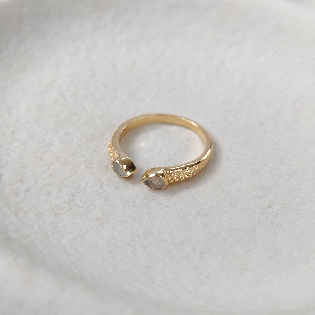Gold ring with a open cuff design and two pear-shaped moonstones on a textured white surface.