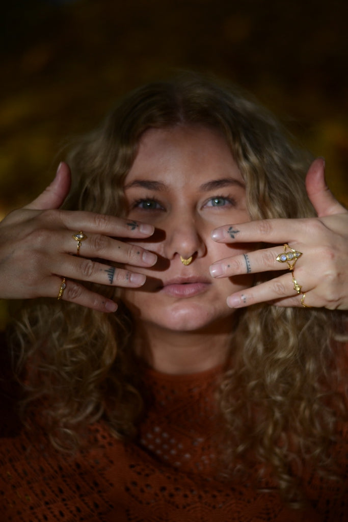 Woman with curly hair holding her hands up to her face, wearing multiple gold  rings with moonstone.