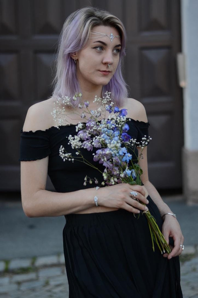 Woman in a black off-shoulder dress, with a moonstone and silver head chain, holding a bouquet of flowers against a dark background