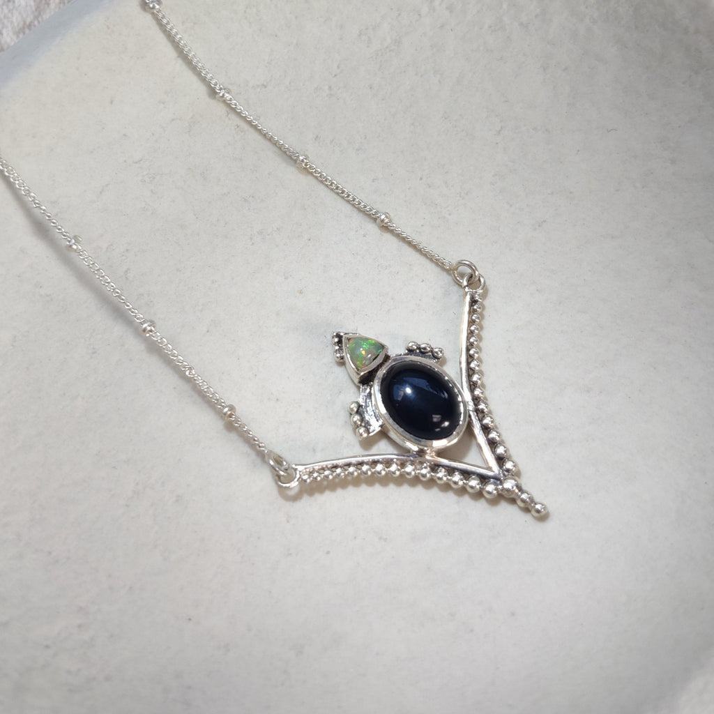 V-shaped silver pendant necklace with black onyx and opal, displayed on a white jewelry plate.