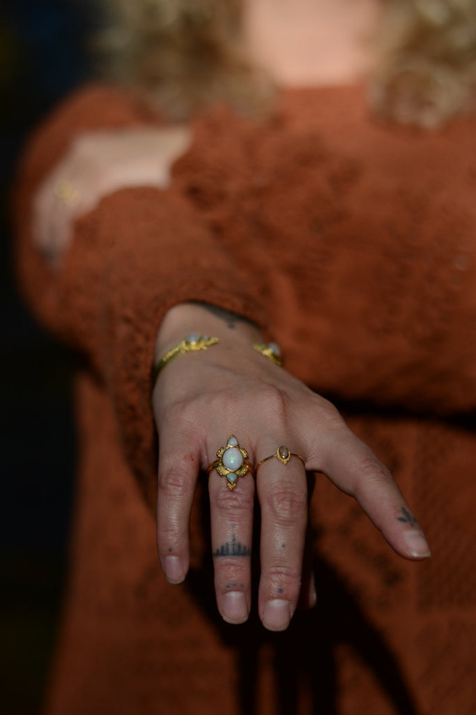 Close-up of a hand with gold rings with opal and tattoos wearing a brown garment.