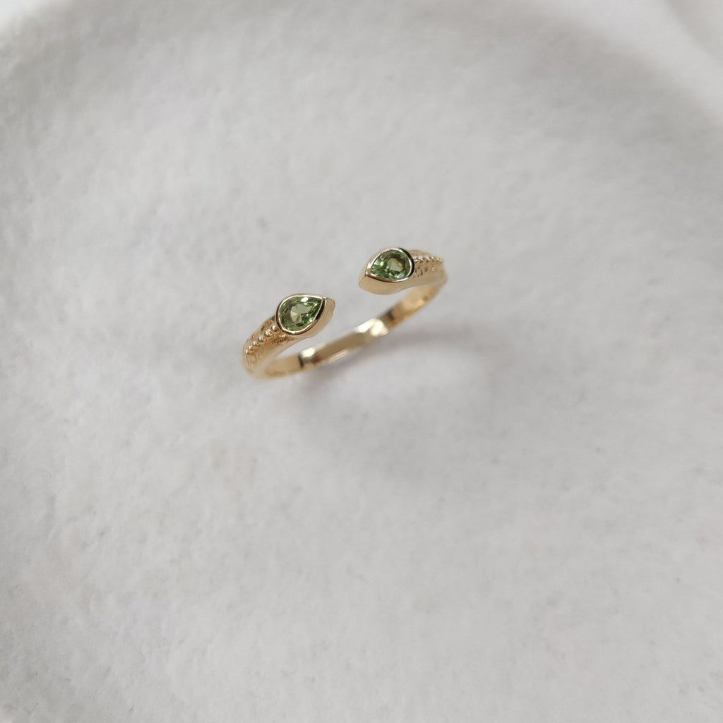 Open cuff ring in gold with green gemstones on a white textured surface.