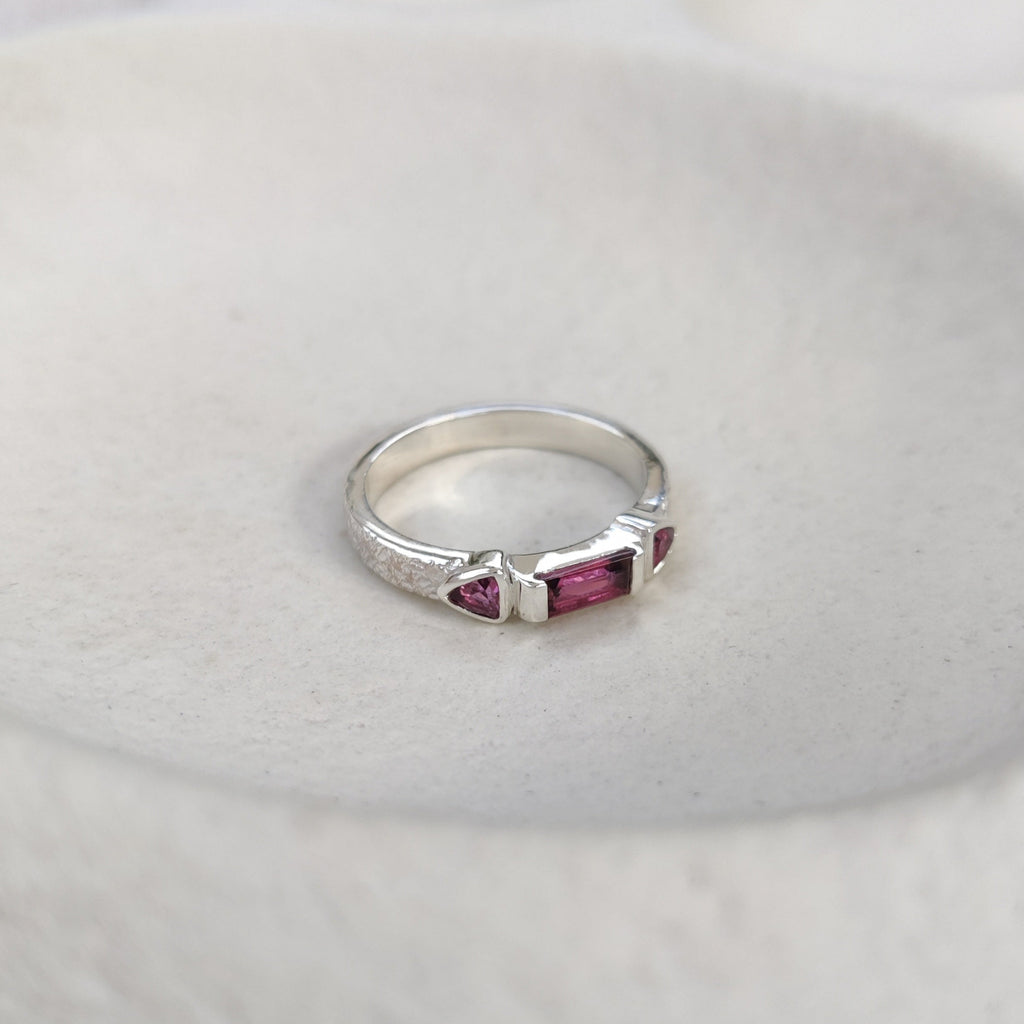 Silver ring with three pink gemstones on a textured white surface.
