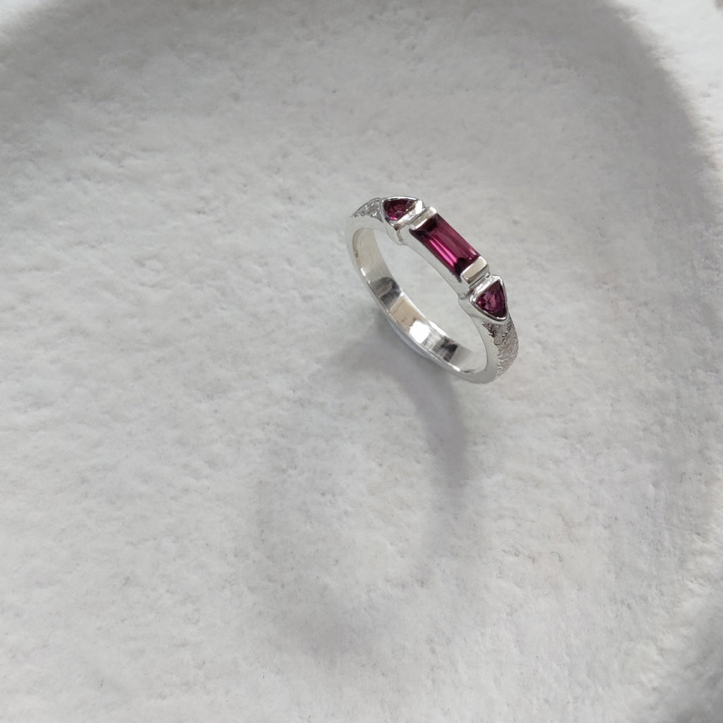 Silver ring with pink gemstones on a textured white surface.