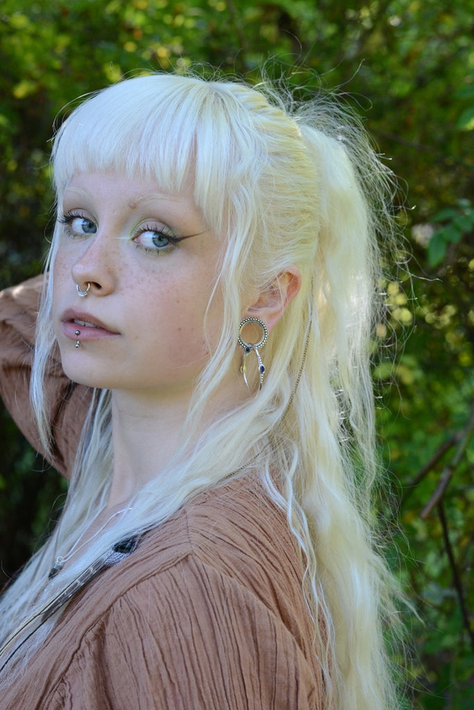 Person with long blonde hair styled in a ponytail, with septum nose ring, tunnels and hoop earrings, wearing a brown dress outdoors.