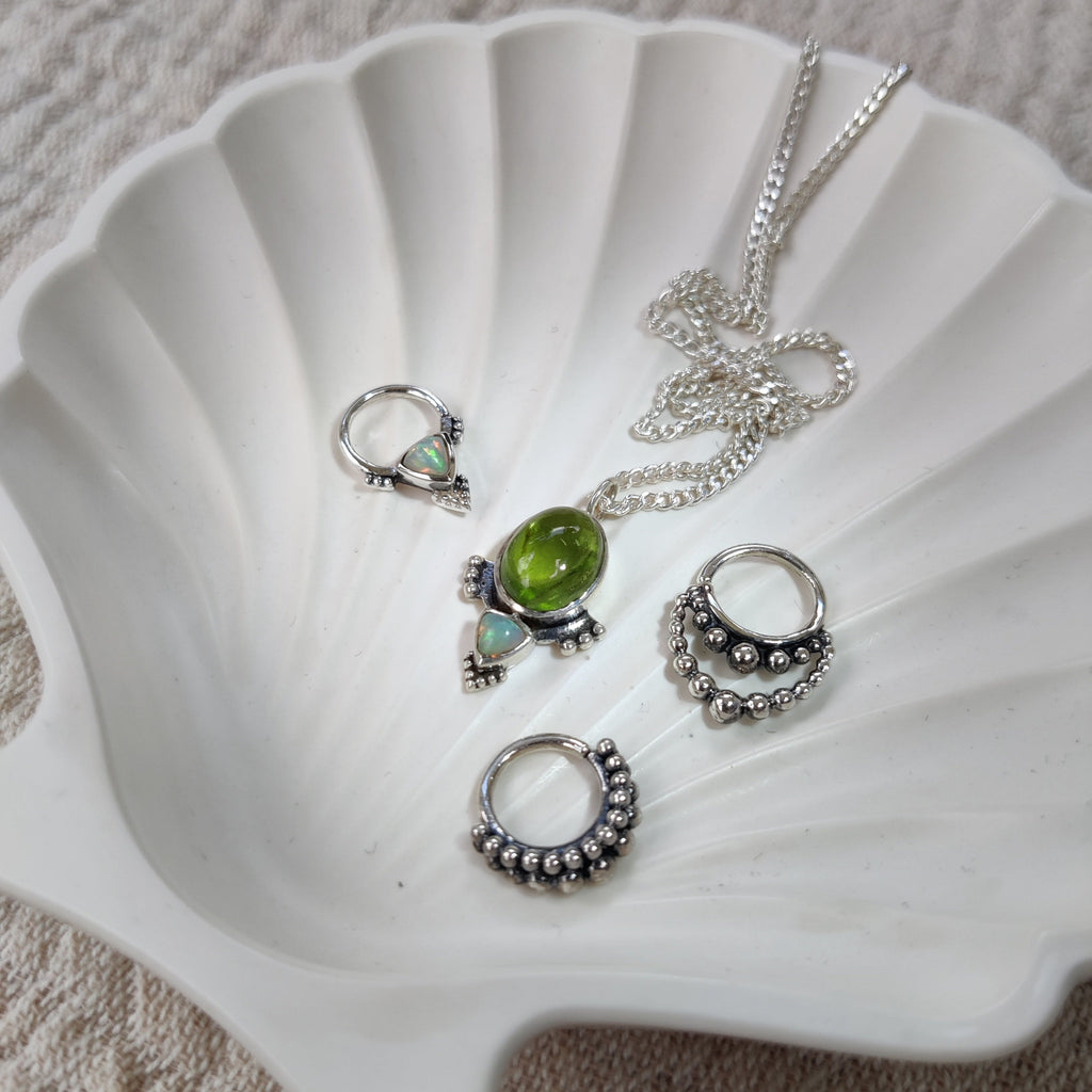 A collection of silver septum jewelry featuring intricate dotted details and opal, and a matching necklace with peridot and opal, displayed on a jewelry dish.