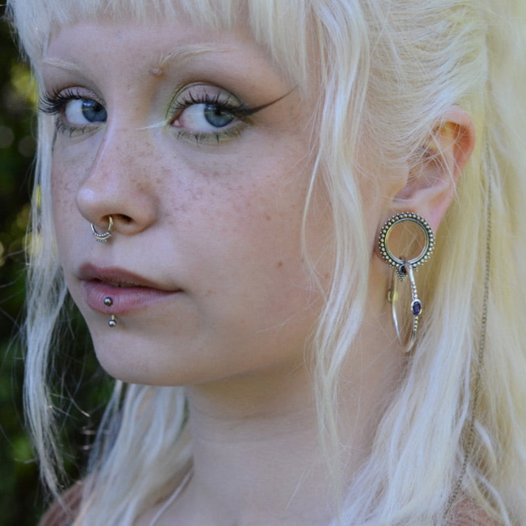 Person with blonde hair and piercings, wearing tunnels and medium hoop earrings against a blurred natural background